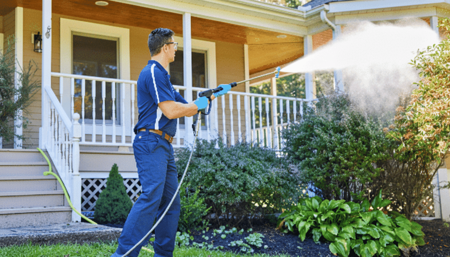 A person uses a pressure washer to clean the exterior of a house, spraying water onto the front porch area near shrubs and a garden.