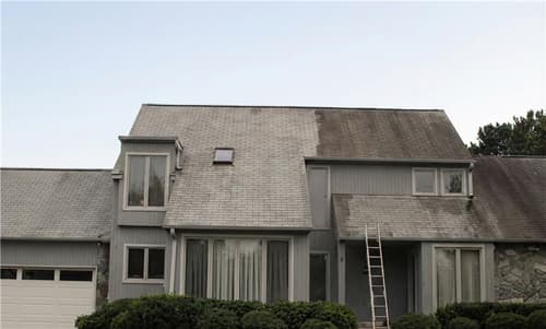 A two-story gray house with a sloped roof and a ladder against the side. There are large windows and greenery in front of the house. The sky appears overcast.