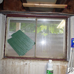 A basement window with duct tape covering one broken pane.  The other pane has been covered up with a wood panel.  A hole has been cut in the panel so a dryer vent could be run through it.