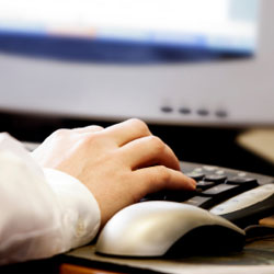 A hand operating a keyboard, with a mouse nearby and the corner of a computer monitor in view.