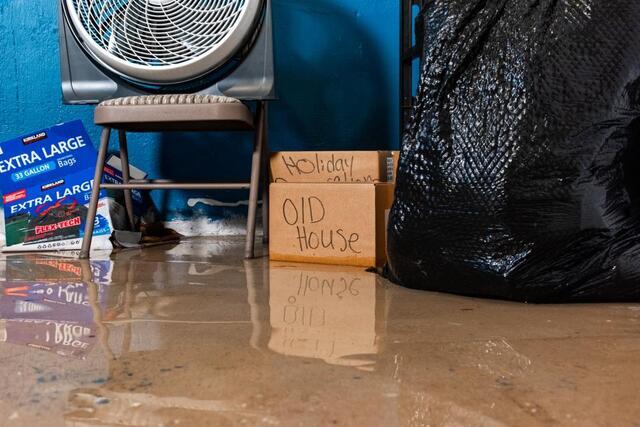 A flooded basement. There is standing water on the ground. Toward the wall, there are some boxes that are wet on the bottom. The closest one reads "old house". There is also a large trash bag and a chair with a large fan on it..