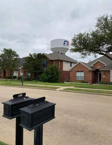 This image captures a quiet residential neighborhood in Allen, Texas. In the foreground, there are two black mailboxes mounted on posts by the curb. Across the street, you see well-maintained brick houses with neatly trimmed lawns and mature trees. Rising prominently in the background is the City of Allen water tower, easily identified by its large white structure and the city&rsquo;s logo featuring a blue and red star. The sky is overcast, giving the scene a calm, subdued atmosphere. This photo highlights the suburban charm and community feel of Allen, TX.