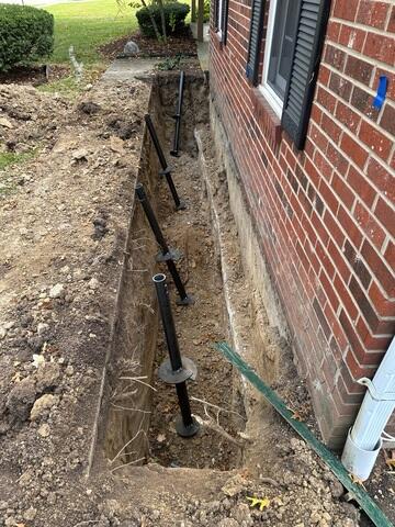The first step in this process was digging a trench to expose the footing of the structure. The footing is the lowest part of a foundation and is typically poured first during a home's construction. In this photo, you can also see our helical piers. Their screw-like design allows them to be drilled deep into the ground with minimal soil displacement, creating a strong, stable connection to the load-bearing strata below.