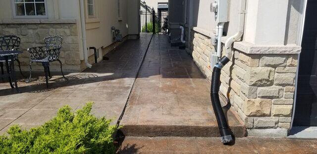 A long, narrow stamped concrete side walkway running between two homes. This view highlights the successful lift and level, showing a clean, even path leading from the patio toward the gated side yard. The newly applied premium crack and joint sealant creates a sharp, professional line between the slabs.