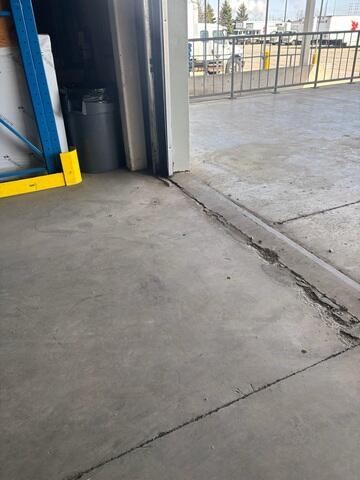 Severe spalling and jagged cracks at the bay door transition, creating a major trip hazard and a jarring "thump" for forklifts.