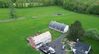 This aerial shot captures the barn mid-installation, with the VeloraONE underlayment and SealoronXT Ice & Water Barrier fully in place along all edges and around the windows. The crew is installing IKO Dynasty shingles in Granite Black, giving the barn a sleek, durable new look. In the background, you can see the second barn's roof with heavy weathering streaks running down, showing the aged condition of the roof.