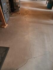 Severe cracking across the concrete slab, visible unevenness, and clear settlement near the home's essential mechanical equipment (furnace/utility area).