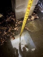 Standing water is visible along the basement wall-floor joint, with a measuring tape placed at the entry point to show the depth of intrusion during active seepage.